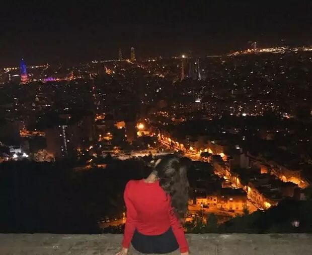 A person with long, dark, curly hair, wearing a red long-sleeved shirt and a black skirt, sits on a ledge overlooking a sprawling cityscape at night. The city lights twinkle below, creating a vibrant and expansive view.
