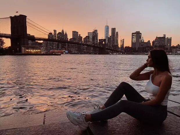A woman sits by the water, gazing at the New York City skyline during sunset, with the Brooklyn Bridge visible in the foreground.