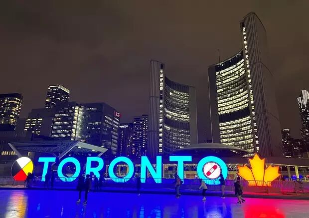 A lit-up TORONTO sign stands before a nighttime skyline, with skaters on an ice rink in the foreground and Toronto City Hall in the background.