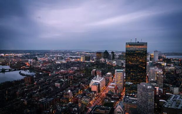 This is a high-angle, dusk-time photograph of the Boston cityscape, showcasing numerous illuminated buildings and a winding river under a cloudy sky.