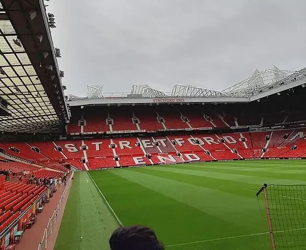 This is a wide-angle shot of the interior of a football stadium, likely Old Trafford, with red seats and green turf visible under a cloudy sky. The text "SATAFORD END" is visible on the stadium