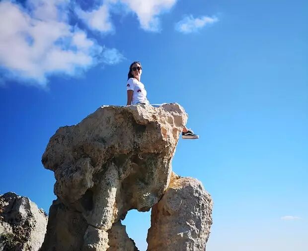 A woman is sitting on a unique rock formation with a hole in it, under a bright blue sky with fluffy white clouds. She is wearing sunglasses and a white t-shirt.