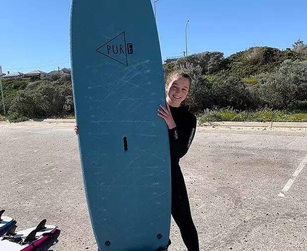 A young woman in a wetsuit smiles as she holds a teal surfboard, ready for a day of surfing. The background shows a sunny day with some vegetation and a parking lot.