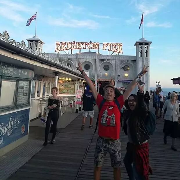 This image shows a lively scene at Brighton Pier, with people enjoying themselves on a sunny day. The iconic pier sign is visible, along with food stalls and the British flag.