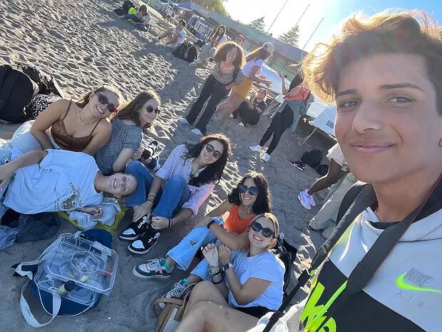 A group of young adults are enjoying a sunny day at the beach, sitting together on the sand and smiling for a selfie.