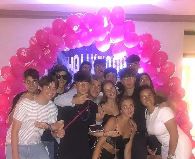 A group of teenagers poses in front of a pink balloon arch and a "HOLLYWOOD" banner, all looking excited and festive.
