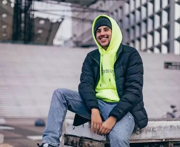 A young man wearing a gray beanie, a neon yellow hoodie, and a black puffer jacket is sitting outdoors, smiling.