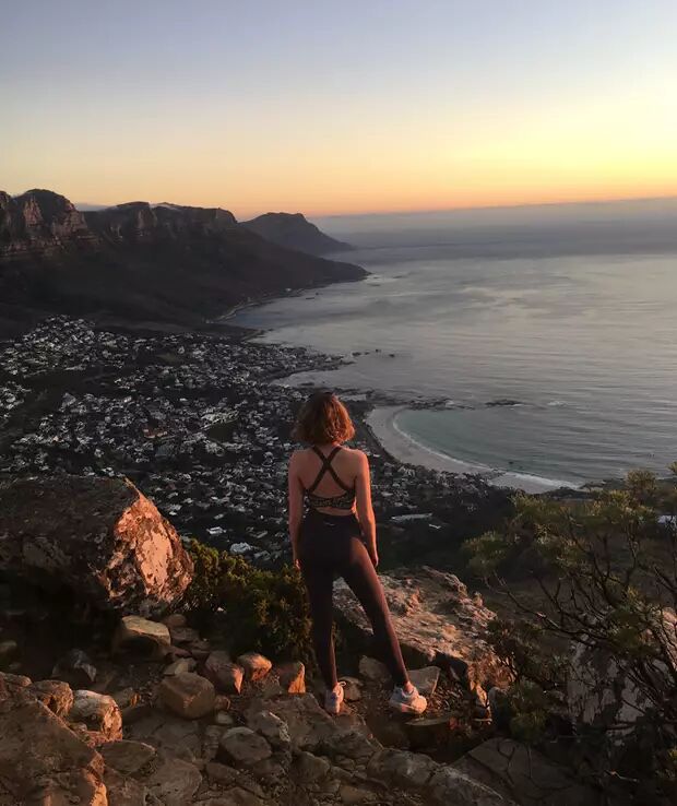 A woman stands on a rocky outcrop, overlooking a city and the ocean during sunset. The view is breathtaking.