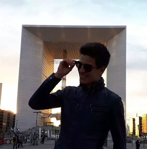 A young man wearing sunglasses and a black jacket adjusts his sunglasses in front of the modern Grande Arche de la Défense in Paris, France.