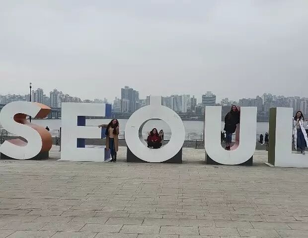 This image features the iconic "SEOUL" sign with three people posing around it. The background shows a cityscape with a river in the foreground, likely a popular tourist spot in Seoul.