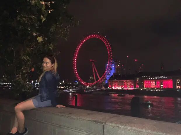 A woman sits on a wall at night with the London Eye illuminated in the background.