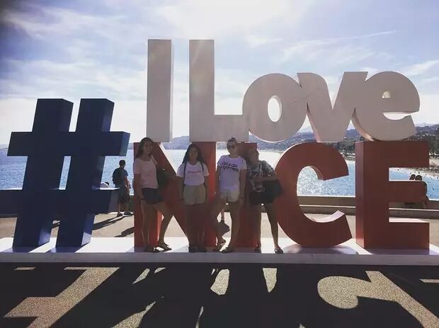 Four young women are happily posing in front of a large "I love Nice" sculpture by the beach, with the blue sea and a sunny sky in the background.