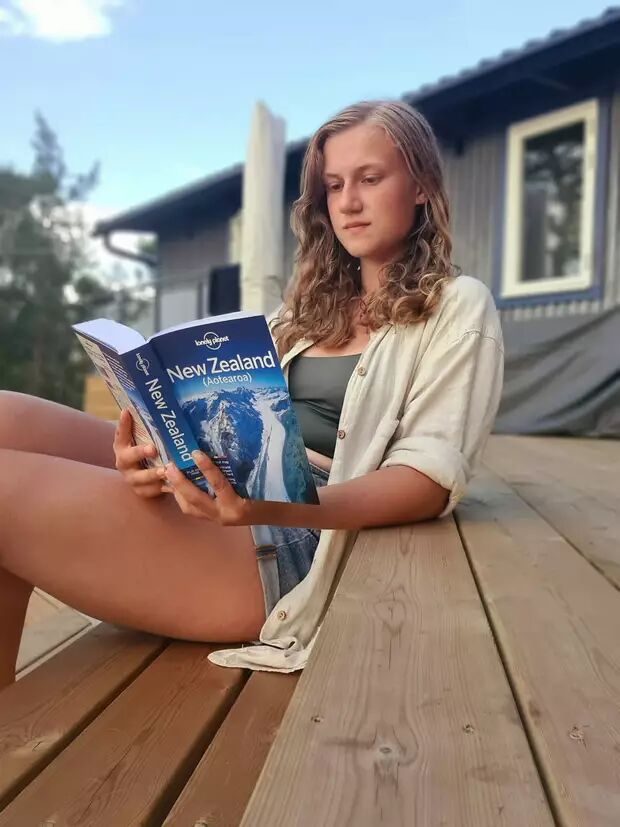 A young woman with long, curly hair is relaxing on a wooden deck, engrossed in reading a travel guide about New Zealand. She is wearing a light-colored shirt over a dark top and denim shorts.