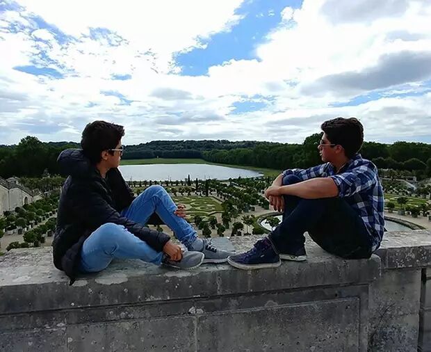 Two young men are sitting on a stone wall, looking at each other, with a beautiful garden and a lake in the background under a cloudy sky.
