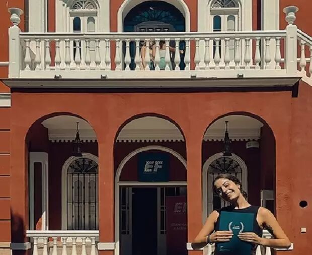 A woman poses in front of a red building featuring arches and "EF" signage. Three other people stand on the balcony above, looking down.