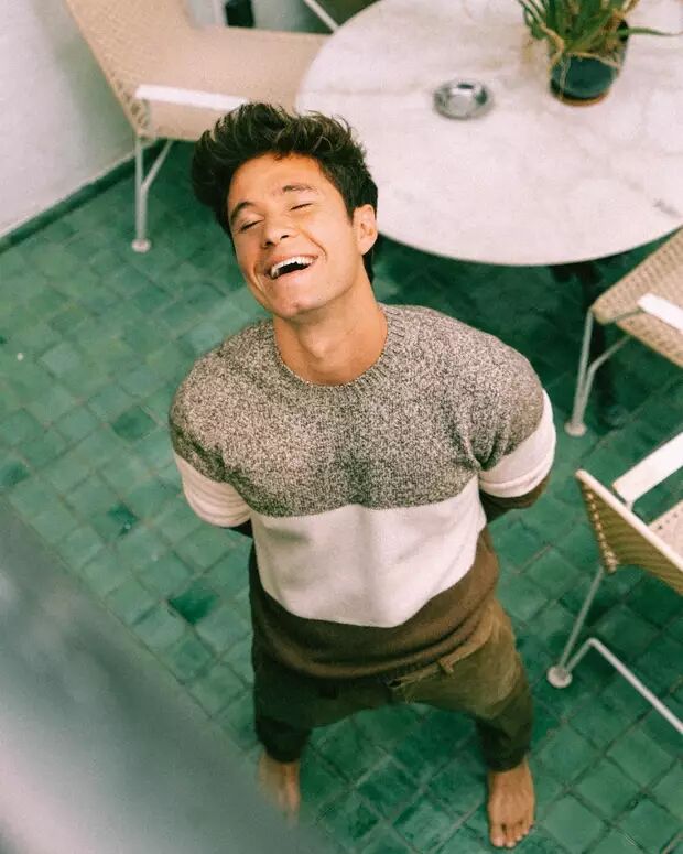 A young man with dark, styled hair is laughing heartily with his eyes closed, wearing a color-blocked sweater and dark pants, standing barefoot on a green tiled floor.