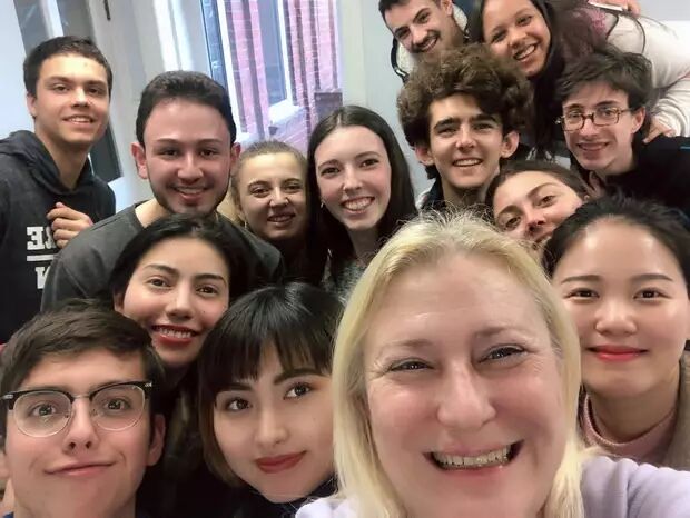 A group of students and a teacher pose for a selfie in a classroom setting. They are all smiling and appear happy.