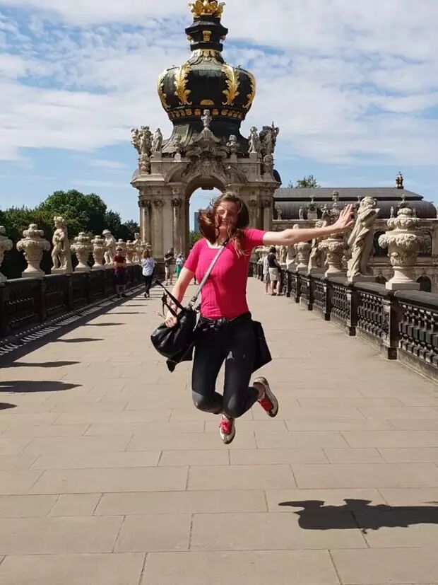 A woman joyfully jumps in the air on a sunny day, with an ornate bridge and a grand architectural structure in the background.
