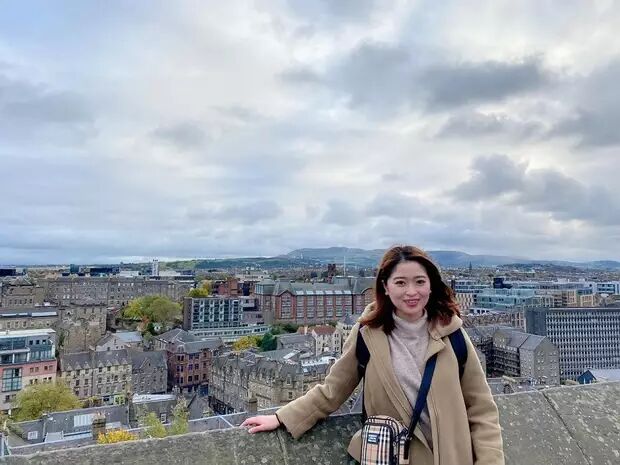 A young woman stands on a rooftop overlooking the city of Edinburgh, Scotland. She is wearing a beige coat and a scarf.