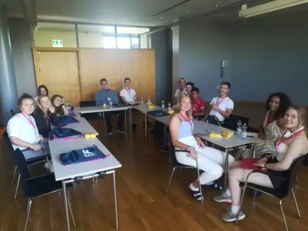 A group of young people, likely students, are seated around a U-shaped table in a conference room, engaged in a discussion or presentation.