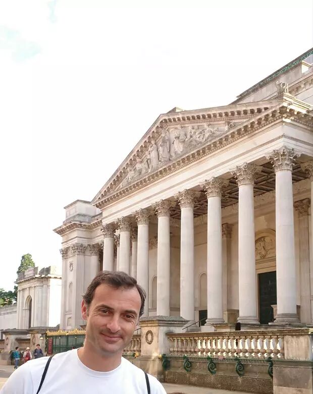 A man in a white t-shirt is standing in front of a large building with columns. He is smiling at the camera.
