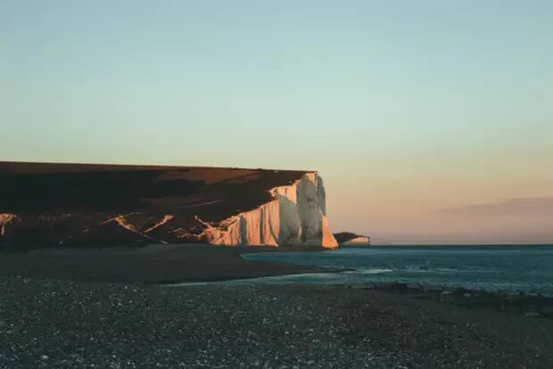The image shows the Seven Sisters chalk cliffs in East Sussex, England, at sunset. The low sun casts a warm glow on the white cliffs, with the sea gently lapping at the pebble beach in the foreground.