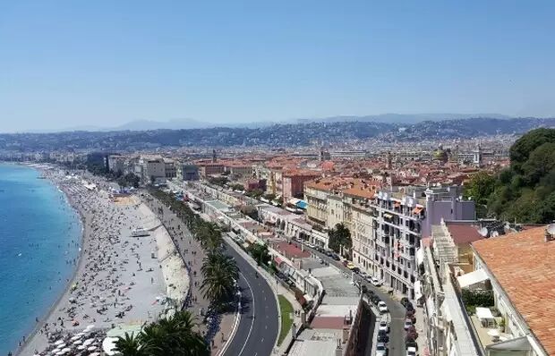 This is a scenic view of the Nice coastline in France, featuring a crowded beach, a palm-lined promenade, and the cityscape extending towards the hills under a clear blue sky.