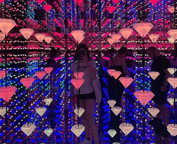 A person stands in a room filled with mirrors and diamond-like lights, creating an infinite reflection. The lighting is predominantly blue and pink.