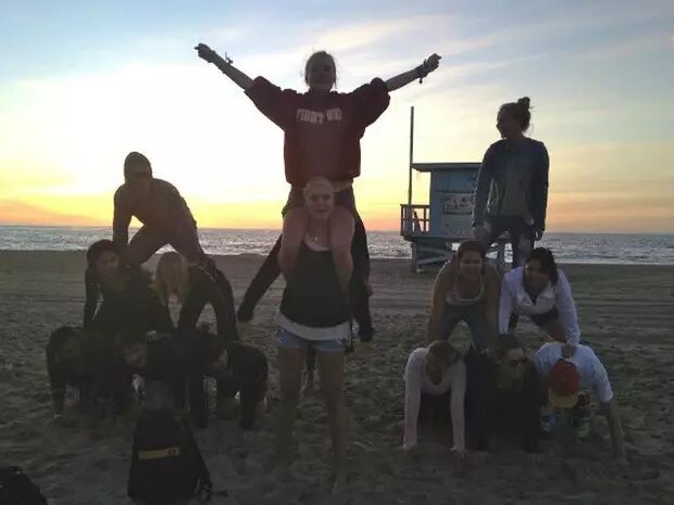 A group of people forms a human pyramid on a beach at sunset, with the ocean and a lifeguard tower in the background.