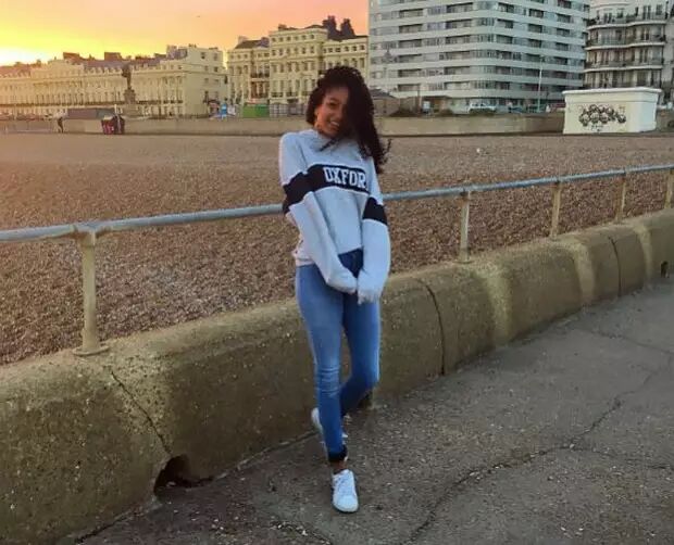 A young woman stands on a beach, smiling at the camera. She is wearing a grey sweatshirt and jeans, with buildings and the ocean in the background.