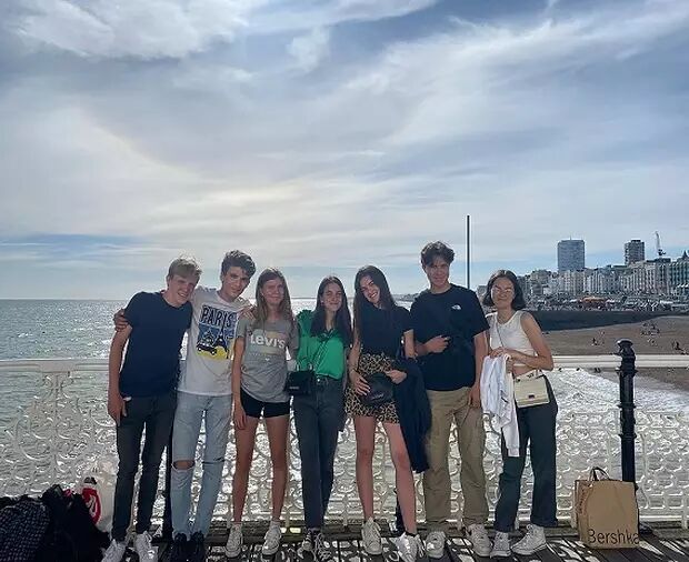 A group of eight young people are posing for a photo on a pier, with a cityscape and cloudy sky in the background. They are casually dressed and appear to be enjoying a day out together.