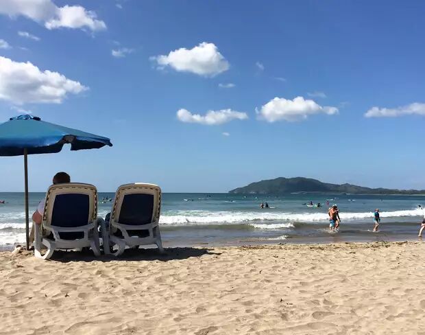 Two lounge chairs sit under a blue umbrella on a sandy beach, overlooking the ocean where people are swimming and surfing.