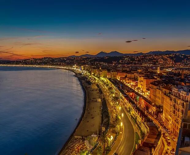 The image shows a beautiful coastal city at dusk, with lights illuminating the beach promenade and buildings. The sea is calm, reflecting the warm glow of the city lights under a darkening sky.