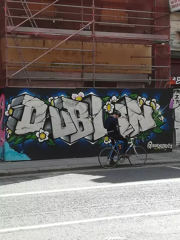 A cyclist rides past a wall featuring graffiti that spells out "DUBLIN." The graffiti is in a 3D style, adorned with flowers and leaves.