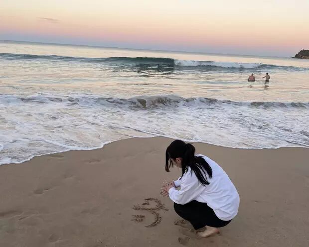 A young woman is drawing in the sand on a beach at sunset, with gentle waves rolling in and a few people in the water.