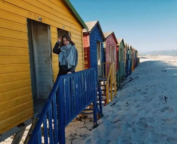 A woman stands in front of a row of colorful beach houses on a sunny day. The wooden huts are painted in bright colors like yellow, blue, and red, and are lined up on the sandy beach.