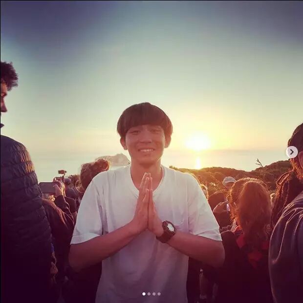 A young man with his hands pressed together in a prayer-like gesture smiles at the camera as the sun sets in the background. He is surrounded by a crowd enjoying a scenic view.
