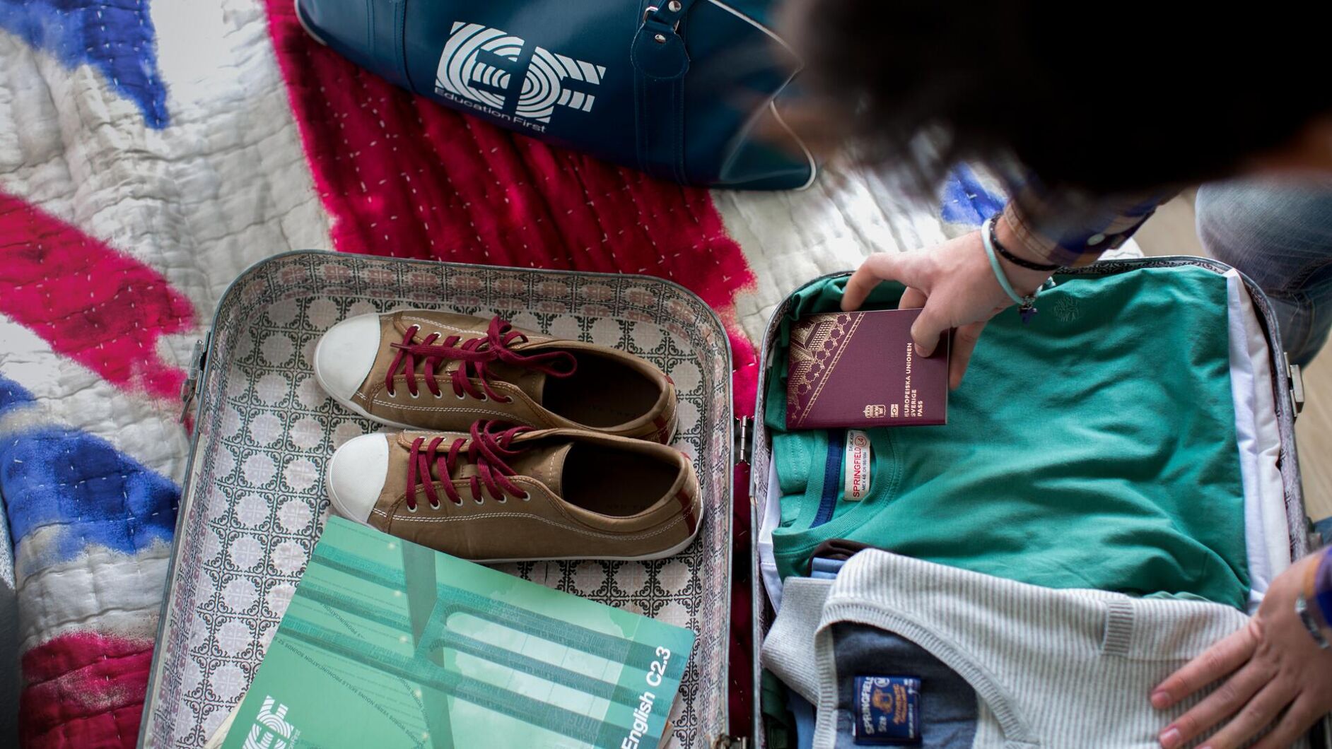 A person is packing a suitcase with clothes, shoes, and a textbook. An EF logo is visible on a bag.