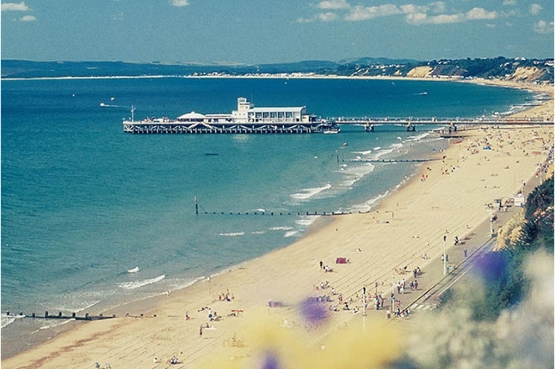 A long pier extends over the calm sea to a sandy beach dotted with people sunbathing and strolling along the shore.