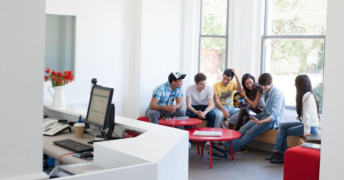 A group of young adults are gathered in a bright room near windows, chatting while they wait.