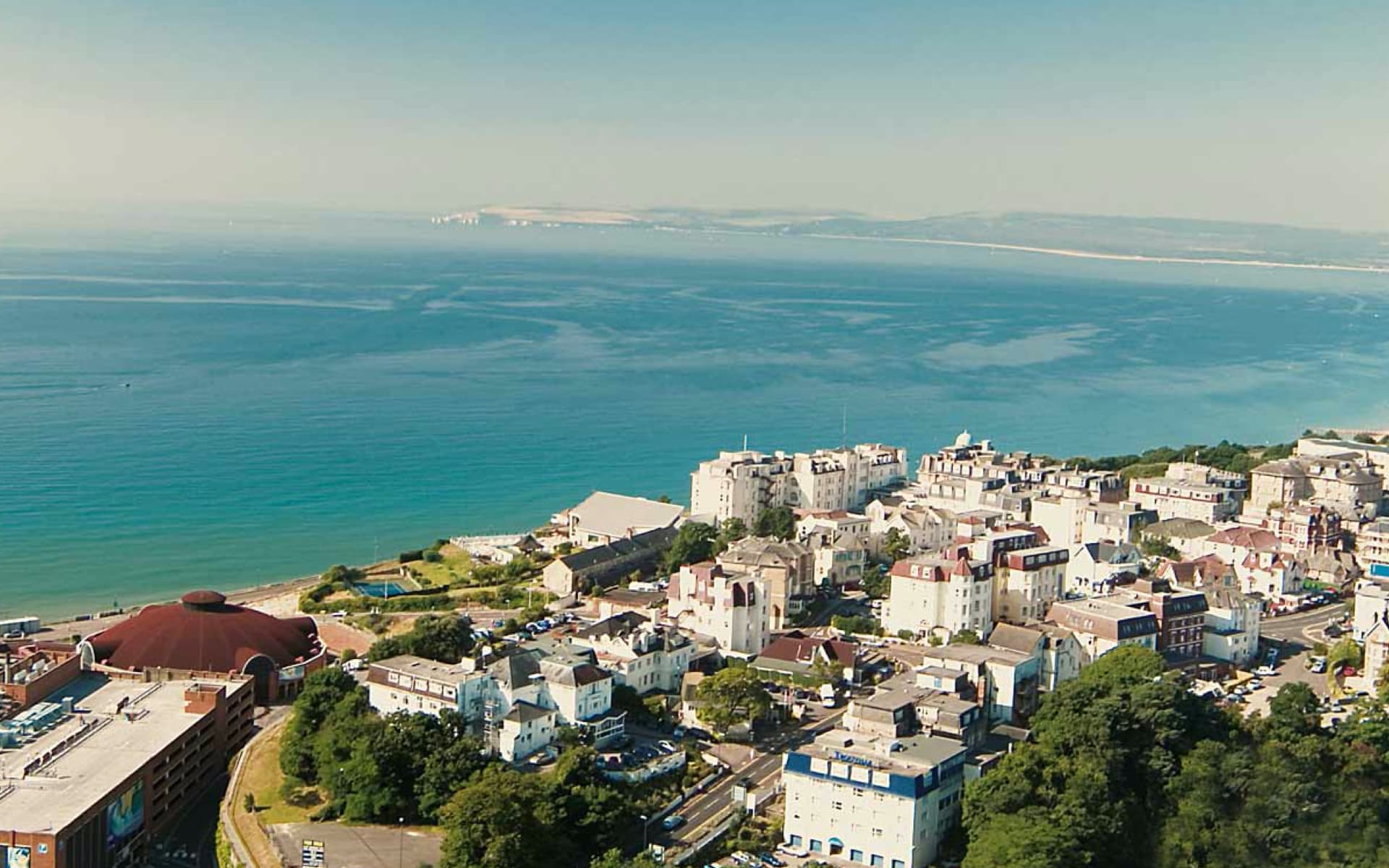 This is a scenic aerial view of a coastal town with white buildings, lush green trees, and the sparkling blue ocean under a clear sky.