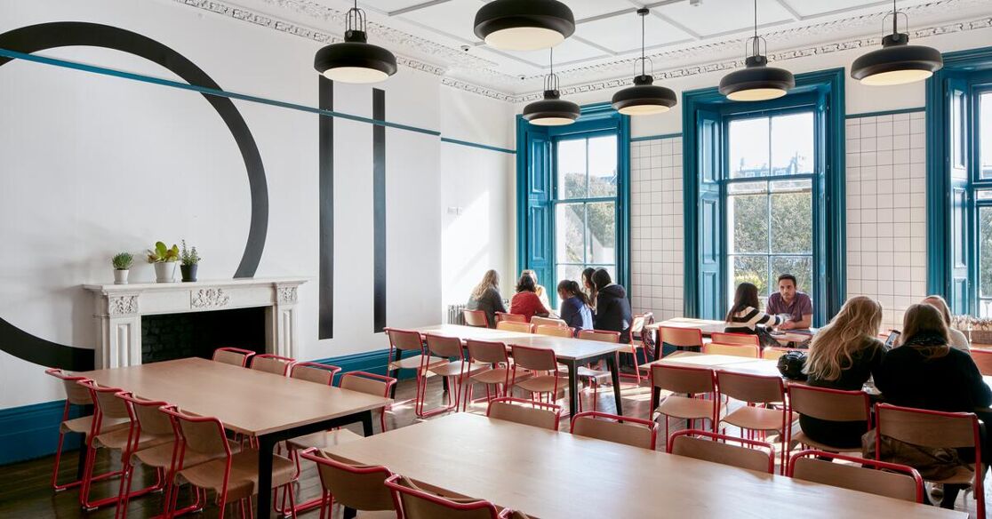 A bright dining hall with long tables and chairs, where several people are conversing. Large windows allow ample daylight.