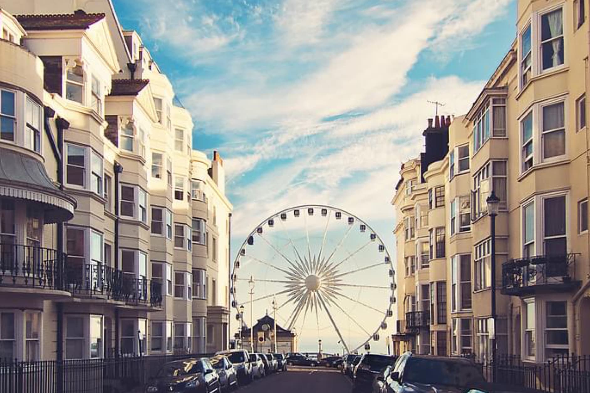 A large Ferris wheel dominates the view down a street lined with Victorian buildings on both sides, with cars parked along the road and a blue sky with clouds above.