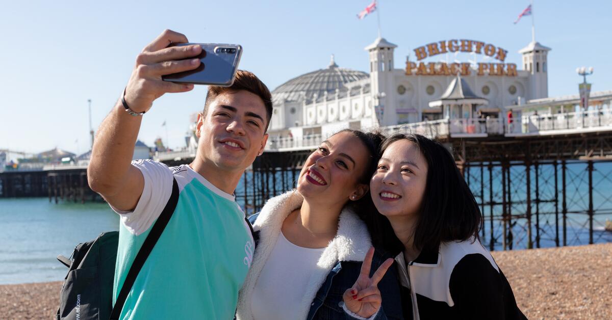 Three young people are taking a selfie on the beach with the Brighton Palace Pier in the background. They are smiling and appear happy.