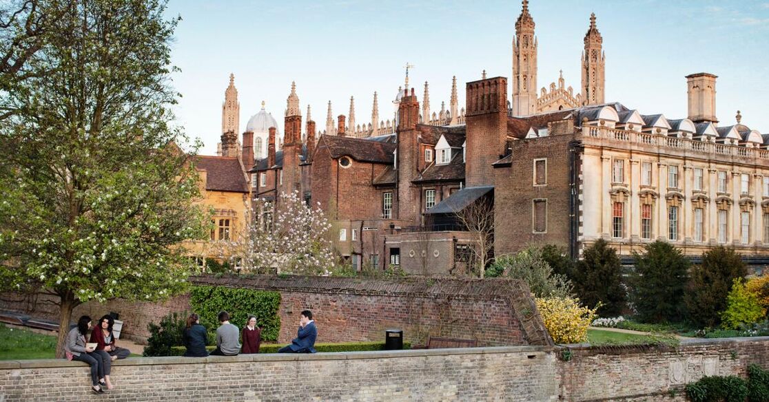 A scenic view of King's College, Cambridge, with its iconic Gothic architecture reflected in the River Cam. Students are seen relaxing by the riverbank on a pleasant day.