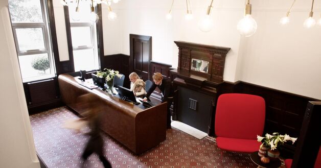 A reception area featuring a wooden desk with three people working, and a red chair beside it. The room has dark wood accents and a patterned carpet tile.