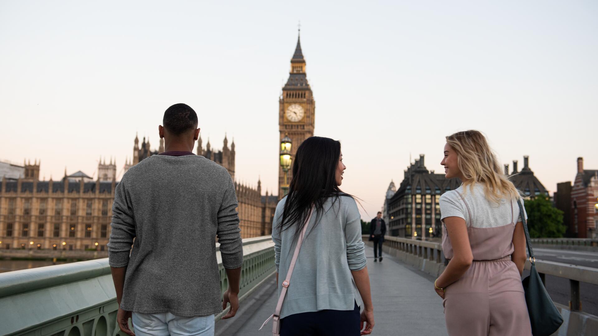 Three friends are walking across a bridge towards the iconic Big Ben clock tower in London, with the Houses of Parliament visible in the background during the golden hour.