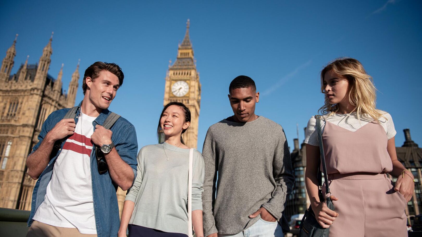 Four young adults are walking outdoors with the iconic Big Ben clock tower in the background, suggesting a tourist excursion in London.