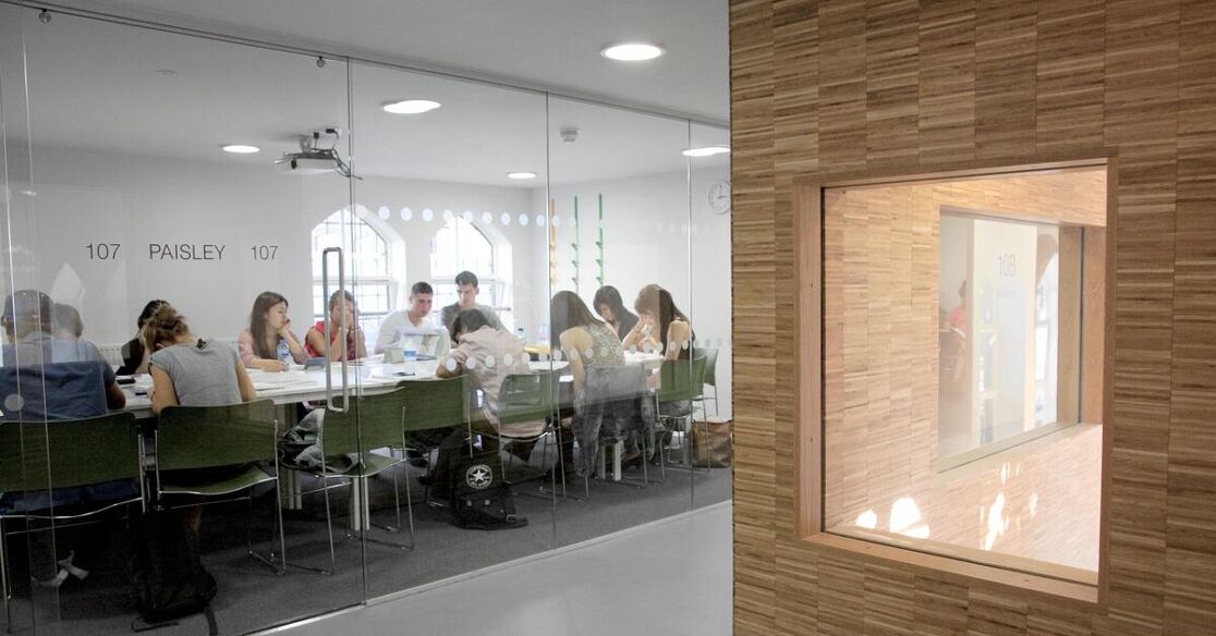 The image shows a group of students in a classroom setting, attentively listening to a lecture or discussion around a conference table. The room features a projector and large windows.