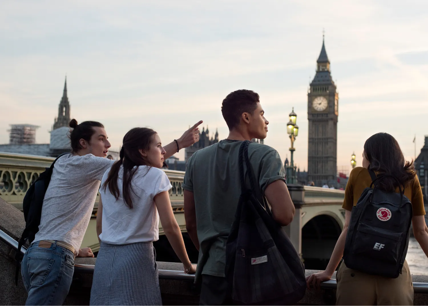 Four young people stand on a bridge, looking out at the London skyline with Big Ben visible in the background. One of them is pointing at something in the distance.
